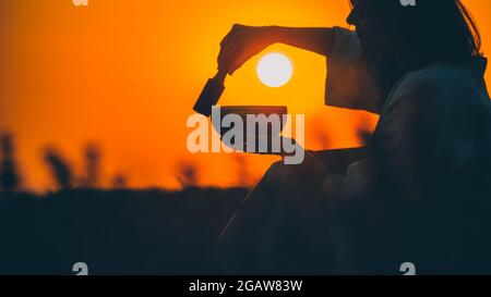 Filmische Silhouette einer Frau, die bei einem wunderschönen orangefarbenen Sonnenuntergang mit Sonnenscheibe eine tibetische Schale spielt. Konzept: Frieden, Ruhe, Zen, Klangheilung Stockfoto