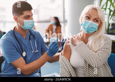 Krankenschwester oder Arzt in medizinischer Gesichtsmaske, die älteren weiblichen Patienten während der Massenimmunisierungskampagne im Gesundheitszentrum Injektionen verabreicht. Ältere Frau ge Stockfoto