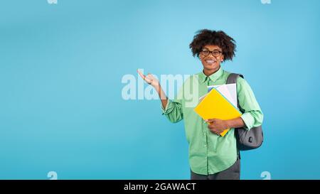 Werbung für Bildungsangebote. Positiver schwarzer Student, der freien Platz für Texte mit Büchern, Notizbüchern und Rucksack zeigt, der über blau steht Stockfoto