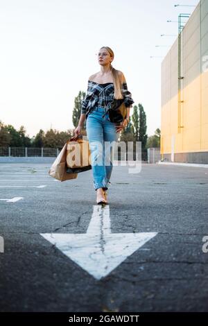 Shopping, Verkauf, Happy Young stilvolle blonde Frau mit Einkaufstaschen gehen zum Auto auf dem Parkplatz auf weißem Pfeil nach dem Einkaufen. Frau mit Einkaufstaschen Stockfoto