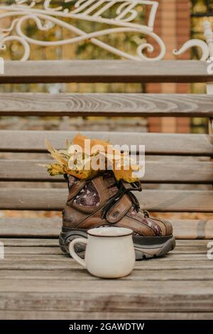 Hallo Herbst, Herbstsaison, Herbststimmung. Gelbe Ahornblätter im Stiefel- und Emaille-Becher auf der Bank im Herbstpark Stockfoto