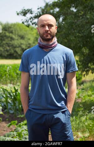 Weißer Mann in einem blauen T-Shirt und Shorts. Ein kahler Mann mit Stoppeln im Gesicht hält seine Hände in seinen Taschen. Blick in die Kamera. Stockfoto