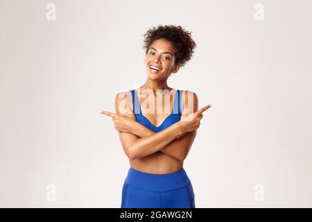 Waist-up Aufnahme der gesunden afroamerikanischen fit Frau in Sportbekleidung für das Training, zeigt die Finger seitlich und lächelnd, zeigt zwei Varianten, weiß Stockfoto