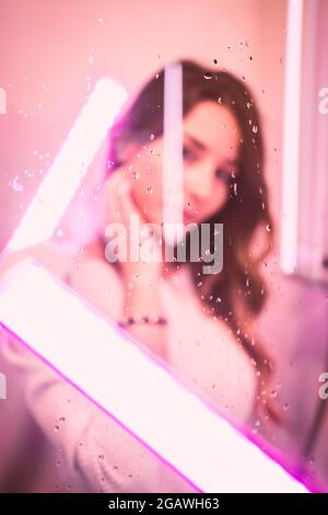 Ein vertikal verschwommenes Porträt einer jungen Frau in rosa Neonlicht durch ein nasses Glas Stockfoto