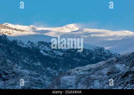 Die Schlucht von Poqueira mit Capileira ragt über den Rand des Berges und der Veleta-Gipfel in der Sierra Nevada im Hintergrund, alles weiß von einem Schnee Stockfoto