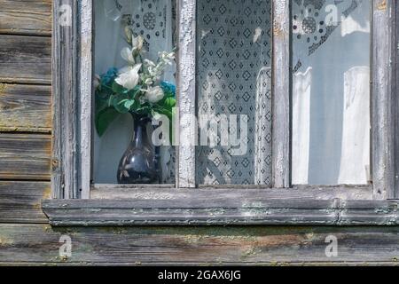 Gealtertes abgeschältes Holzfenster mit weißen traditionellen Vorhängen und einem Blumenstrauß aus synthetischen Blumen in einer blauen handbemalten Keramikvase Stockfoto