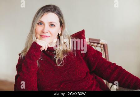 Attraktive Frau mittleren Alters, die am Wochenende auf einem bequemen Stuhl zu Hause sitzt und sich entspannt, trägt einen farbenfrohen, warmen roten Strickpullover und lächelt an Stockfoto