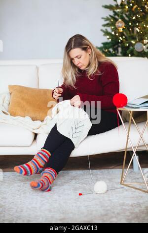 Frau mittleren Alters sitzt auf der Couch im Wohnzimmer Strickdecke mit Mustern mit Nadeln, glückliche Frau zu Hause entspannen, während sie Lieblings-Hobby Stockfoto