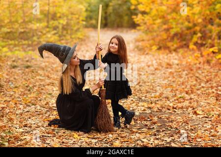 Ganzkörperfrau und Mädchen in Hexenkostümen fegen verwelkte Blätter vom Weg mit Besen während der Halloween-Feier im Herbstwald Stockfoto