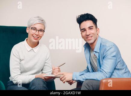 Frau mit Stift und Notizbuch in der Hand schreiben und Mann sitzt auf einem Stuhl zusammen. Vertrag zur Umfrage zu Journalismus, Interviews und Personalbeschaffung Stockfoto
