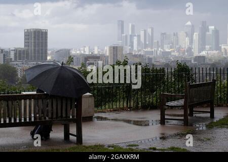 30/07/2021. London, Großbritannien. Ein Mann unterzieht sich unter einem Regenschirm, während er während eines Regensturms auf Point Hill in Greenwich so über die Canary Wharf blickt Stockfoto