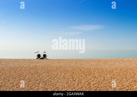 Isoliertes Paar, das unter Sonnenschirmen am Kiesstrand in St. Leonards on Sea, East Sussex, sitzt Stockfoto