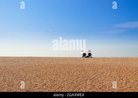Isoliertes Paar, das unter Sonnenschirmen am Kiesstrand in St. Leonards on Sea, East Sussex, sitzt Stockfoto