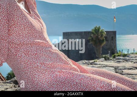 Eine Frau in EINEM roten Kleid sitzt auf der Steinmauer gegen EINE schöne Landschaft mit Schloss in der Mitte. Stockfoto