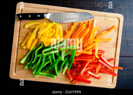Rote, orange, gelbe und grüne Paprika in dünne Streifen geschnitten: Paprika in Schlagstöcke auf einem Bambusschneidebrett mit einem Kochmesser geschnitten Stockfoto