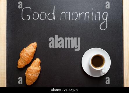 Tasse Kaffee, Croissants, die Aufschrift „Guten Morgen“ auf einer schwarzen Tafel. Ein traditionelles Frühstück, guter Start in den Tag Konzept. Speicherplatz für kopieren Stockfoto