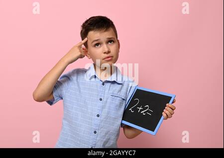 Ein Schuljunge mit einer Tafel in der Hand hält einen Finger an seinen Tempel und löst mit einem nachdenklichen Blick eine Rechenaufgabe, stehend auf einem rosa Backgr Stockfoto