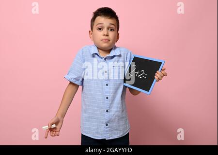 Ein süßer Schuljunge hält eine Tafel in der Hand mit einem Rechenproblem und mit Überraschung und Fragen schaut er auf die Kamera, während er auf einem Rosa steht Stockfoto