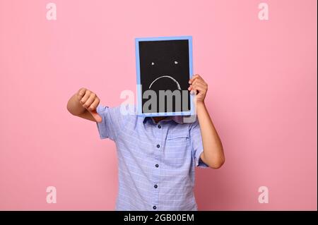 Ein Schuljunge bedeckt sein Gesicht mit einer Tafel mit gezeichneten lächelnden Emoticons, drückt Traurigkeit aus und zeigt den Daumen zur Kamera. Isoliert über Rosa Stockfoto
