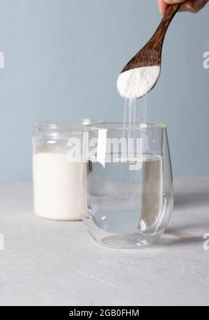 Kollagenpulver wird mit einem Löffel in ein Glas Wasser gegeben. Gesundes Anti-Age-Konzept auf hellblauem Hintergrund. Vertikales Format Stockfoto