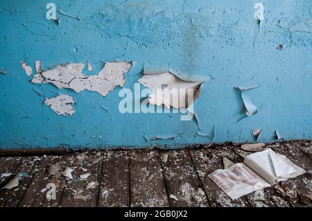 Verlassene Grundschule. Blätternde blaue Farbe an der Wand. Buch liegt auf dem Boden. Stockfoto