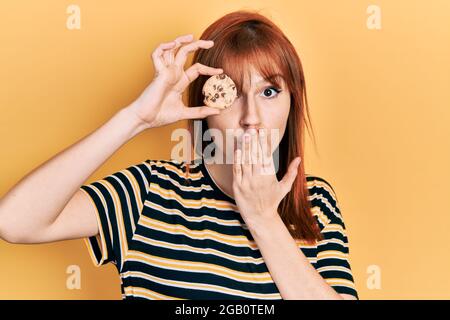 Rotschopf junge Frau hält Cookie in der Nähe Auge bedeckt Mund mit der Hand, schockiert und Angst vor Fehler. Überrascht Ausdruck Stockfoto