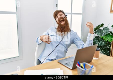 Junger irischer Callcenter-Agent, der lächelnd Musik hört, während er im Büro eine Gitarrengeste macht. Stockfoto