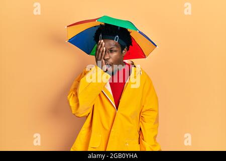 Junger afroamerikanischer Mann mit gelbem Regenmantel, gähnend, müde, bedeckt Gesicht, Auge und Mund mit der Hand. Gesicht schmerzt vor Schmerzen. Stockfoto