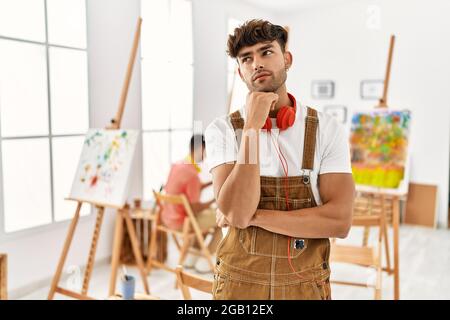 Junger hispanischer Mann im Kunststudio mit Hand auf Kinn, der über Fragen nachdenkt, nachdenklichen Ausdruck. Lächelnd mit nachdenklichen Gesicht. Zweifelskonzept. Stockfoto