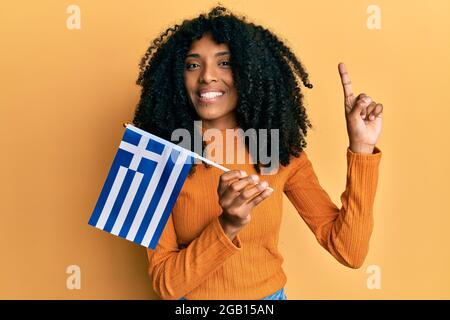 afroamerikanische Frau mit afrohaaren, die die griechische Flagge hält, lächelt mit einer Idee oder Frage, die den Finger mit einem glücklichen Gesicht zeigt, Nummer eins Stockfoto