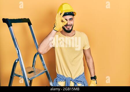 Ein hübscher Mann mit Bart an einer Bautreppe, der einen Hardhut trug, was mit einer Geste mit lächelnder Hand in Ordnung war, ein Auge, das durch die Finger mit einem glücklichen Gesicht schaute. Stockfoto