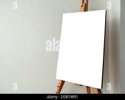 Fotoleinwand auf Staffelei. Weiße leere Kunsttafel auf hölzerner Staffelei stehen auf weißem Wandhintergrund in der Ecke des Künstlerstudios mit Kopierraum. Stockfoto