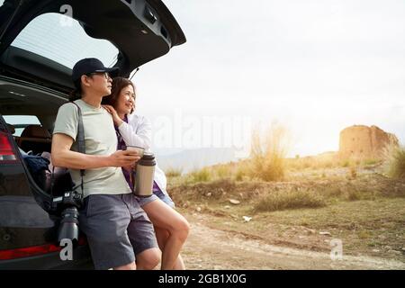 das asiatische Paar lehnte sich an der Rückseite des Autos und schaute auf die Aussicht, während es Kaffee an einem verlassenen historischen Ort trank Stockfoto