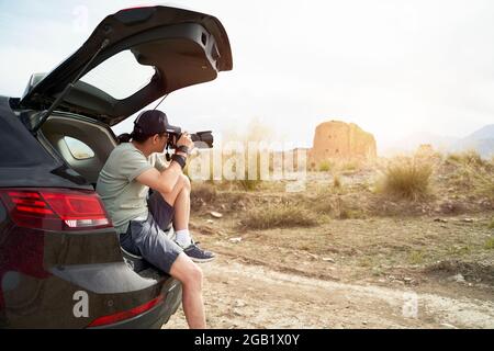 asiatischer Fotograf, der am Kofferraum eines Autos sitzt und ein Bild von einer verlassenen Burg an einem historischen Ort fotografiert Stockfoto