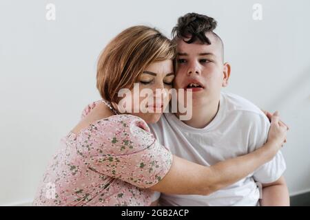 Mutter umarmt behinderten Jungen 17 Jahre alt im Rollstuhl mit zerebraler Lähmung auf weißem Hintergrund. Frau hilft Person mit besonderen Bedürfnissen. Emotional Stockfoto