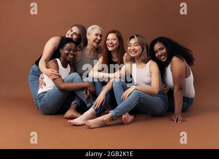 Porträt von sechs lachenden Frauen unterschiedlichen Alters und Körpertypen, die auf braunem Hintergrund im Studio sitzen Stockfoto