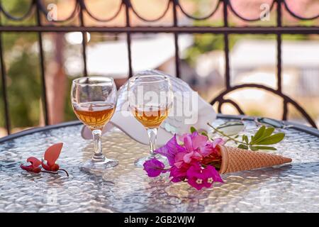 Sommercocktails mit rosa Blüten von Bougainvillea auf einem Glastisch. Erfrischungskonzept. Sommer helle surreale Blumen, alkoholische Liköre und Frauen Sommerhut Stockfoto