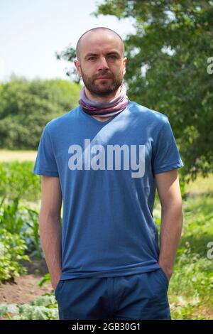 Weißer Mann in einem blauen T-Shirt und Shorts. Ein kahler Mann mit Stoppeln im Gesicht hält seine Hände in seinen Taschen. Blick in die Kamera. Stockfoto