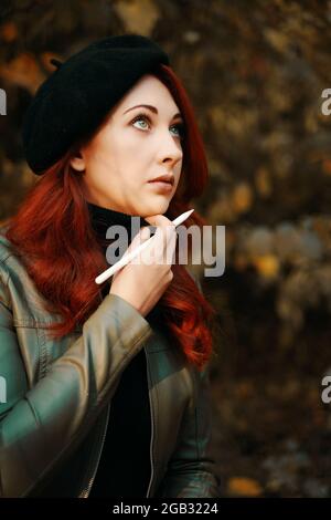 Brütende rothaarige Frau im Park. Hübsche Frau in einer Baskenmütze und Lederjacke. Mann mit Stift, Designer, Freiberufler. Herbstporträt. Warten auf Inspiration. Stockfoto