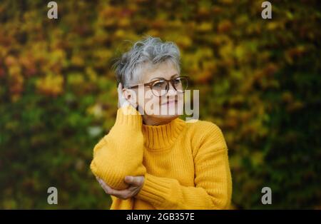 Ältere Frau, die im Freien vor farbenfrohem, natürlichem Herbsthintergrund steht und sich ausruht. Stockfoto