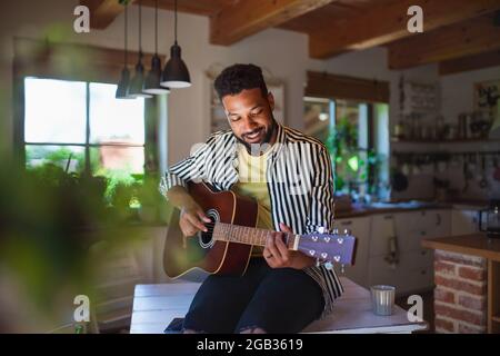 Glücklicher junger Mann mit Gitarre drinnen zu Hause, entspannend. Stockfoto