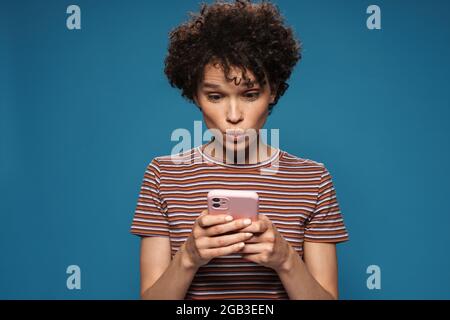 Junge lockige überrascht Frau in gestreiftem T-Shirt mit Mobiltelefon isoliert auf blauem Hintergrund Stockfoto