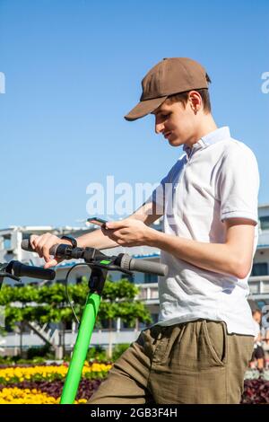Ein junger Mann mietet sich einen Elektroroller, indem er den QR-Code mit einem Smartphone scannt. Scooter auf dem Parkplatz teilen. Stockfoto