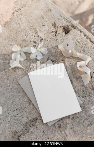 Sommerhochzeit Schreibwaren Nachbau. Leere Grußkarte, Einladung mit Bastelumschlag, Seidenband. Weiße Oleander Blüten auf beigem Beton Stockfoto