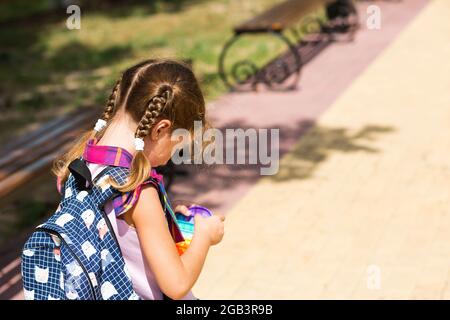 Kleines Mädchen mit Rucksack und in einer Schuluniform im Schulhof spielt Pop-it-Spielzeug. Zurück zur Schule, 1. September. Der Schüler entspannt sich nach dem Unterricht. Stockfoto