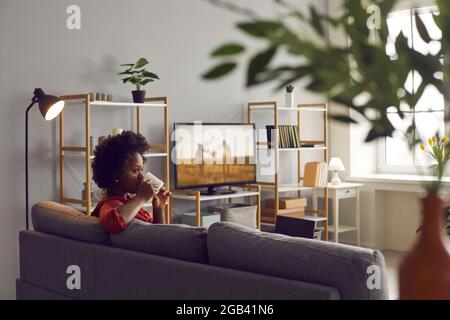 Schwarze Frau sitzt auf dem Sofa im Wohnzimmer, trinkt Tee und schaut sich einen Film im Fernsehen an Stockfoto