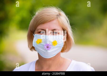 Nahaufnahme Porträt einer Frau im Freien trägt medizinische Gesichtsmaske mit Blumen kümmert sich um ihren Körper im Sommer in Pandemie Zeit. Stockfoto