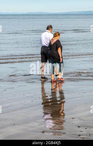 Ein Paar mittleren Alters, das an einem Sandstrand spazieren geht, ein älteres Paar, das am Strand spazieren geht, ein Rentnerpaar, das einen entspannten Spaziergang am Sandstrand macht. Stockfoto