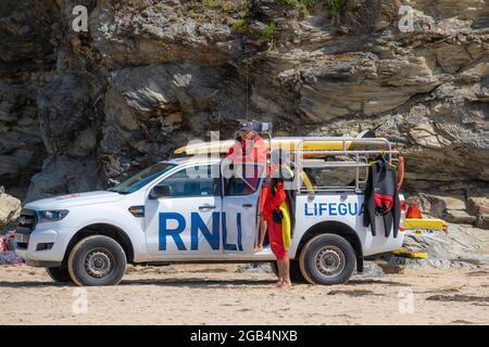 Zwei RNLI-Rettungsschwimmer plaudern am Mawgan Porth Beach in Cornwall mit einem Rettungsfahrzeug, das bei sonnigem Wetter am Strand in Großbritannien geparkt ist. Stockfoto