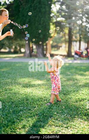 Frau bläst Seifenblasen und kleines Mädchen fangen sie auf einer Lichtung Stockfoto